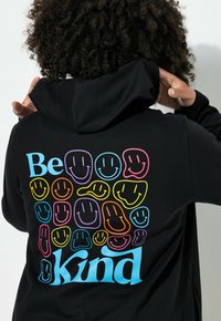 Persona con cabello rizado que lleva puesta una sudadera negra con caras sonrientes coloridas y el texto "Sé amable" en la parte de atrás.