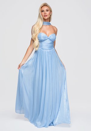 Femme blonde portant une robe fluide bleu clair sans bretelles avec un foulard assorti autour du cou, tenant les côtés de la robe sur un fond uni.
