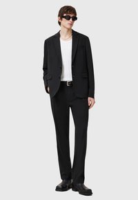 Svart kavaj och byxor, vit ribbad skjorta, svarta skor, svarta rektangulära solglasögon, med en figursydd design och rena linjer.