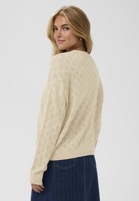 Pull en maille beige clair avec un motif de petits trous, coupe décontractée, manches longues et col rond, associé à un jean en denim foncé.