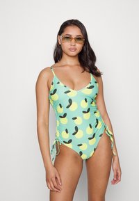 Fato de banho verde com padrão de frutas amarelas, franzido lateral ajustável e alças finas. A modelo usa óculos de sol amarelos e tem cabelo ondulado.