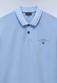 Lys blå piké skjorte med teksturert stoff, to svarte knapper og brodert logo med flaggdesign på venstre brystområde.
