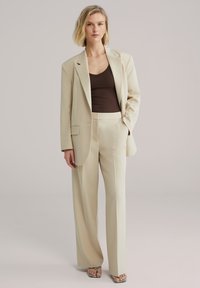 Béžové sako s voľným strihom, v kombinácii s béžovými nohavicami s širokými nohavicami. Pod tým priliehavý tmavohnedý tielko. Doplnky zahŕňajú vzorované sandále.