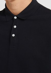 Černé polo tričko s texturovanou tkaninou, s dvoubarevným zapínáním na knoflíky a embosovaným logem nad levou stranou.