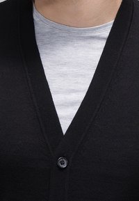 Svart stickad cardigan med V-ringning, prydd med en ensam svart knapp. Bärs över en ljusgrå t-shirt med rundad hals.