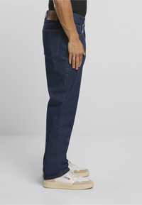 Mies, joka pukeutunut tummansinisiin suoralahkeisiin farkkuihin ja valkoisiin lenkkareihin, seisoo sivuttain käsi reisillä lepäämässä.