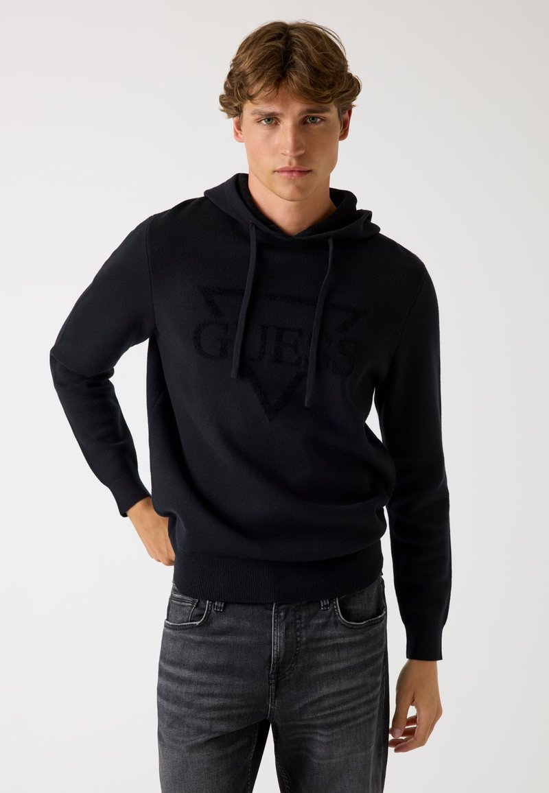 Felpa nera con cappuccio realizzata in tessuto morbido, che presenta un logo "GUESS" testurizzato in un design triangolare sulla parte anteriore, abbinata a jeans grigi.
