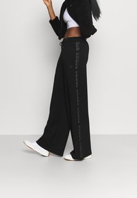 Svarta byxor med vida ben i ett mjukt material, med dragsko i midjan och ett vertikalt logomönster längs sidan. Matchas med vita skor.