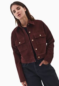 Brązowa kurtka z kordurowej tkaniny, krótka, z dwiema kieszeniami na piersi, złotymi zatrzaskami i wywijanym kołnierzem. Stylizowana z czarną bluzką i ciemnymi spodniami.