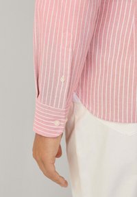 Camicia a righe rosa chiaro con linee verticali bianche. Presenta un polsino con un bottone e un orlo leggermente curvo. Tessuto a trama uniforme.