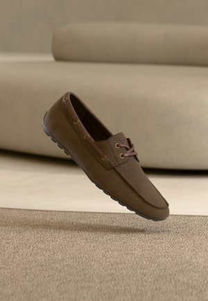 Pantof mocasin din piele maro cu detalii de șiret, talpă de cauciuc, plutind deasupra unei covor texturat într-un ambient interior în tonuri neutre.