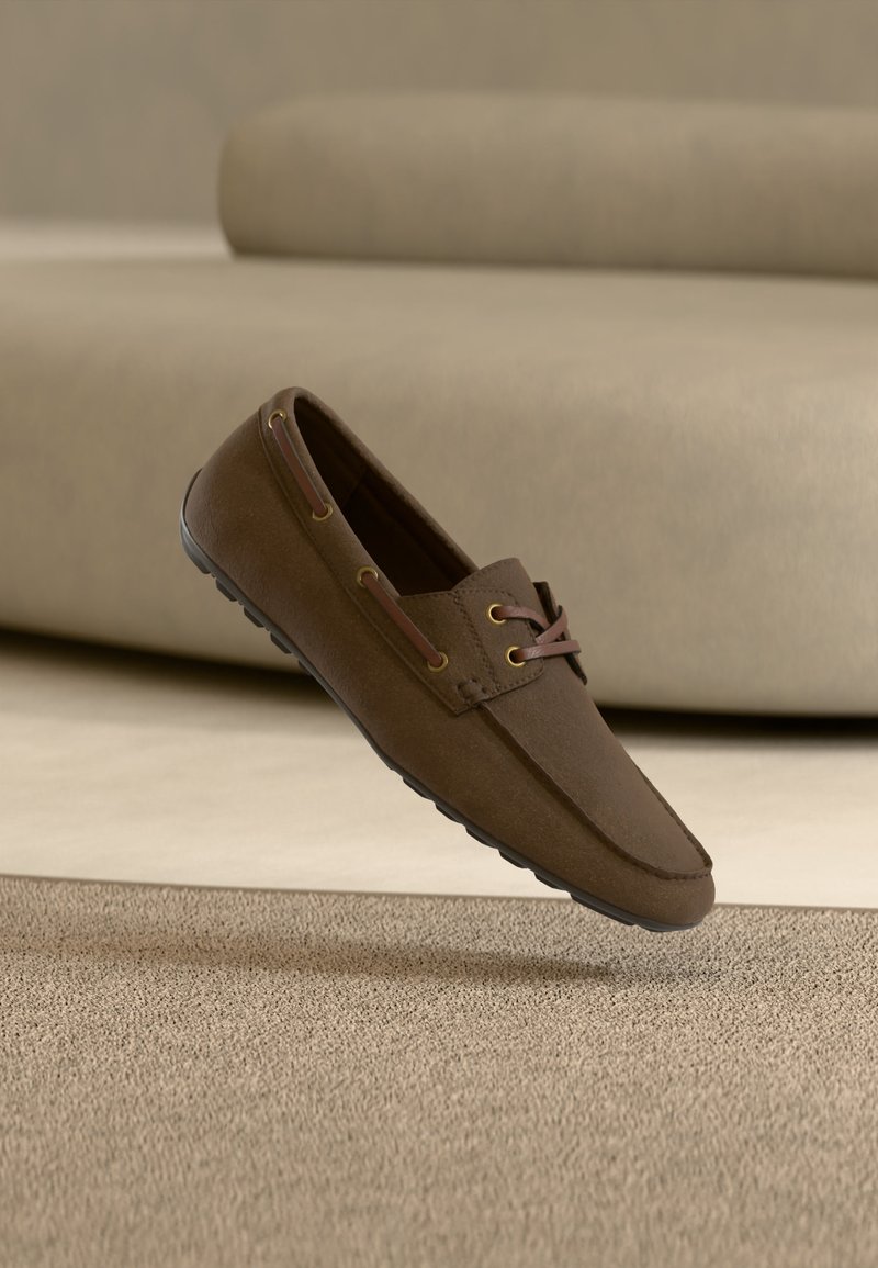 Καφέ μοκασίνι από δέρμα με λεπτομέρειες κορδονιών, ελαστική σόλα, αιωρούμενο πάνω από υφασμένο χαλί σε εσωτερικό χώρο με ουδέτερο χρωματικό τόνο.