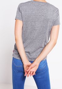 Osoba w szarej koszulce z krótkim rękawem i niebieskich dżinsach stoi z rękami złożonymi za plecami, odwrócona plecami do kamery.