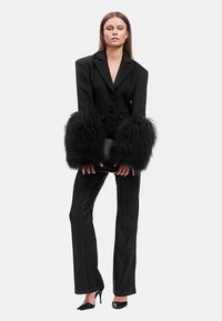 Blazer negro a medida con puños de pelo sintético extragrande, combinado con pantalones negros de corte recto y tacones de punta afilada. Un elegante bolso de mano completa el conjunto.