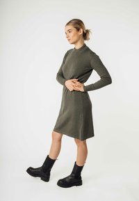 Olivenfarbendes Strickkleid mit hohem Kragen und langen Ärmeln, das eine lockere Passform und einen knie-langen Saum hat. Kombiniert mit schwarzen Ankle Boots.