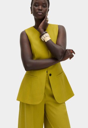 Vrouw in een mouwloze mosterdgele broekpak met wijde pijpen, geaccentueerd met grote armbanden en een ring, poserend met gekruiste armen.