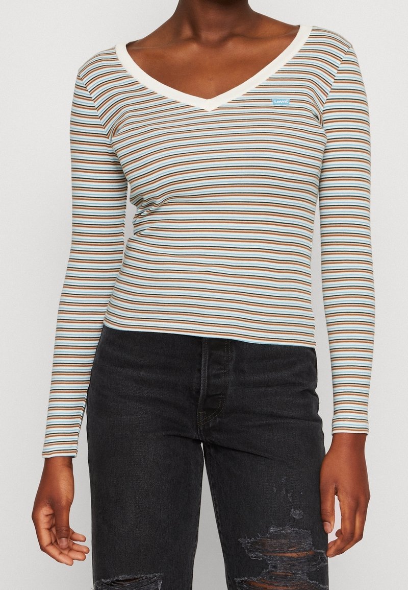 Langärmliges gestreiftes Oberteil in Blau, Braun und Creme, mit V-Ausschnitt und einem kleinen Logo-Tag. Kombiniert mit schwarzen, kaputten Jeans.