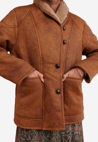 Bruine schapenleren jas met grote knopen en zakken, gedragen over een bloemenpatroon rok, handen in de zakken gestoken.