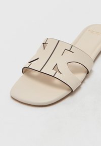 Sandal en toile beige avec une large sangle découpée au design géométrique et un bord noir. Texture lisse et semelle plate.