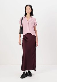 Gestreiftes pinkes Kurzarmshirt kombiniert mit einem burgunderfarbenen Satin-Maxikleid. Das Model trägt schwarze klobige Schuhe und eine schwarze Handtasche.