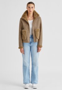 Braune Jacke mit Kunstfellkragen und Taschen, mit Reißverschluss vorne, weiche Textur. Kombiniert mit hellblauen Jeans und weißen Sneakern. Minimalistisches Design.