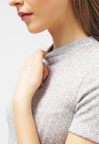 Jasnoszary prążkowany top z krótkimi rękawami i wysokim kołnierzem, wykonany z fakturowanego materiału. Ręka delikatnie trzyma obszar kołnierza.