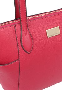 Sac fourre-tout en cuir rouge texturé avec une plaque dorée, des poignées solides et des détails de couture contrastants. Compartiment zippé sur le dessus.