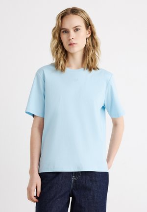 Junge Frau mit welligem blondem Haar, die ein hellblaues T-Shirt mit kurzen Ärmeln und dunkelblaue Jeans vor einem schlichten weißen Hintergrund trägt.