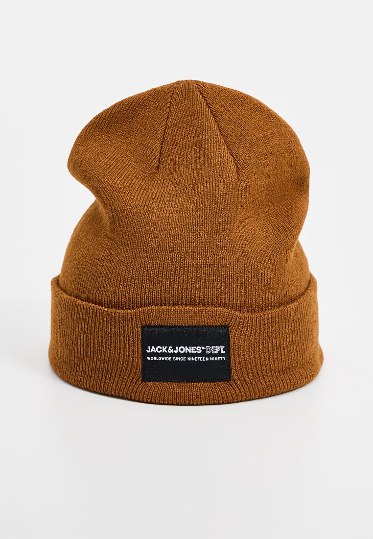 Bruine gebreide beanie muts met een omgeslagen rand. Heeft een zwart label aan de voorkant met witte tekst. Zachte structuur en eenvoudig ontwerp.