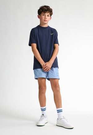 Granatowy bawełniany t-shirt z zielonym logo, w parze z jasnoniebieskimi szortami i białymi trampkami z zielonymi akcentami oraz skarpetkami w niebieskie paski.