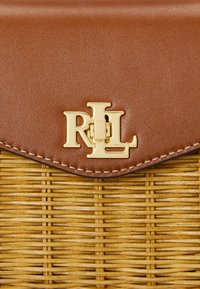 Καφέ δερμάτινη καπάκι με χρυσό λογότυπο "RL", πάνω από ύφασμα σε κίτρινη απόχρωση. Διακριτή ραφή κατά μήκος της άκρης. Συμπαγής σχεδίαση με υφή.