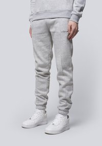 Light grey sweatpants with ribbed cuffs, side stitching details, and a black embroidered logo on the thigh, paired with white sneakers.