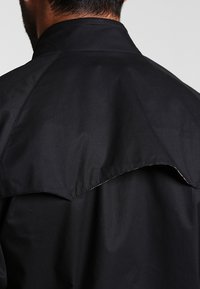Giubbotto nero con colletto alto e una distintiva cucitura a punta sul retro. La trama è liscia, realizzata in un tessuto leggero.