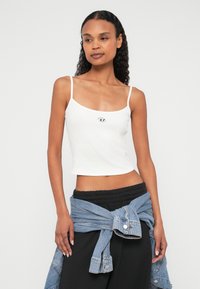 Λευκό κοντό τοπ με λεπτές τιράντες, με μικρό μαύρο λογότυπο στο κέντρο. Συνδυασμένο με μαύρο παντελόνι και τζιν μπουφάν δεμένο στη μέση.