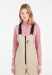 Beige overall met een ritssluiting aan de voorkant, voorzien van verstelbare zwarte bretels. Draag een lichtroze longsleeve top met een hoge kraag eronder.