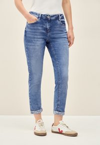 Blå denimjeans med smal passform, uppvikta ärmar, framfickor och knappstängning. I kombination med vita sneakers med röda detaljer.