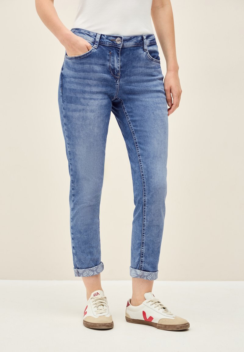 Blå denimjeans med smal passform, uppvikta ärmar, framfickor och knappstängning. I kombination med vita sneakers med röda detaljer.