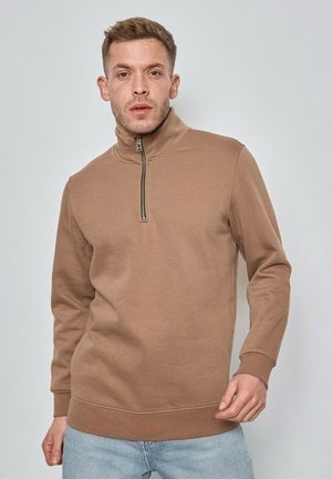 Sweatshirt beige à demi-zippé en tissu doux, avec un col montant, des poignets côtelés et une coupe décontractée, porté avec un jean bleu clair.