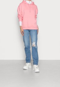 Sudadera con capucha de color rosa claro, con un bolsillo frontal, llevada sobre una camiseta de manga larga blanca; combinada con unos vaqueros rotos de color azul claro y zapatillas blancas.