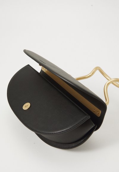 Melnas ādas crossbody soma ar atvērtu vāku, zelta pogu aizdari un viļņotu zelta metāla siksnu uz baltā fona.