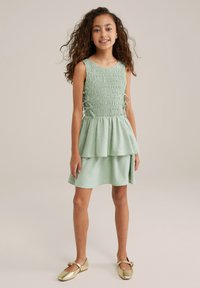 Mėtų žalios spalvos be rankovių suknelė su smock'd detalėmis, sluoksniuota sijonu ir tekstūruotu audiniu su subtiliais raštais. Dėvima su auksinėmis plokščiomis bateliais.