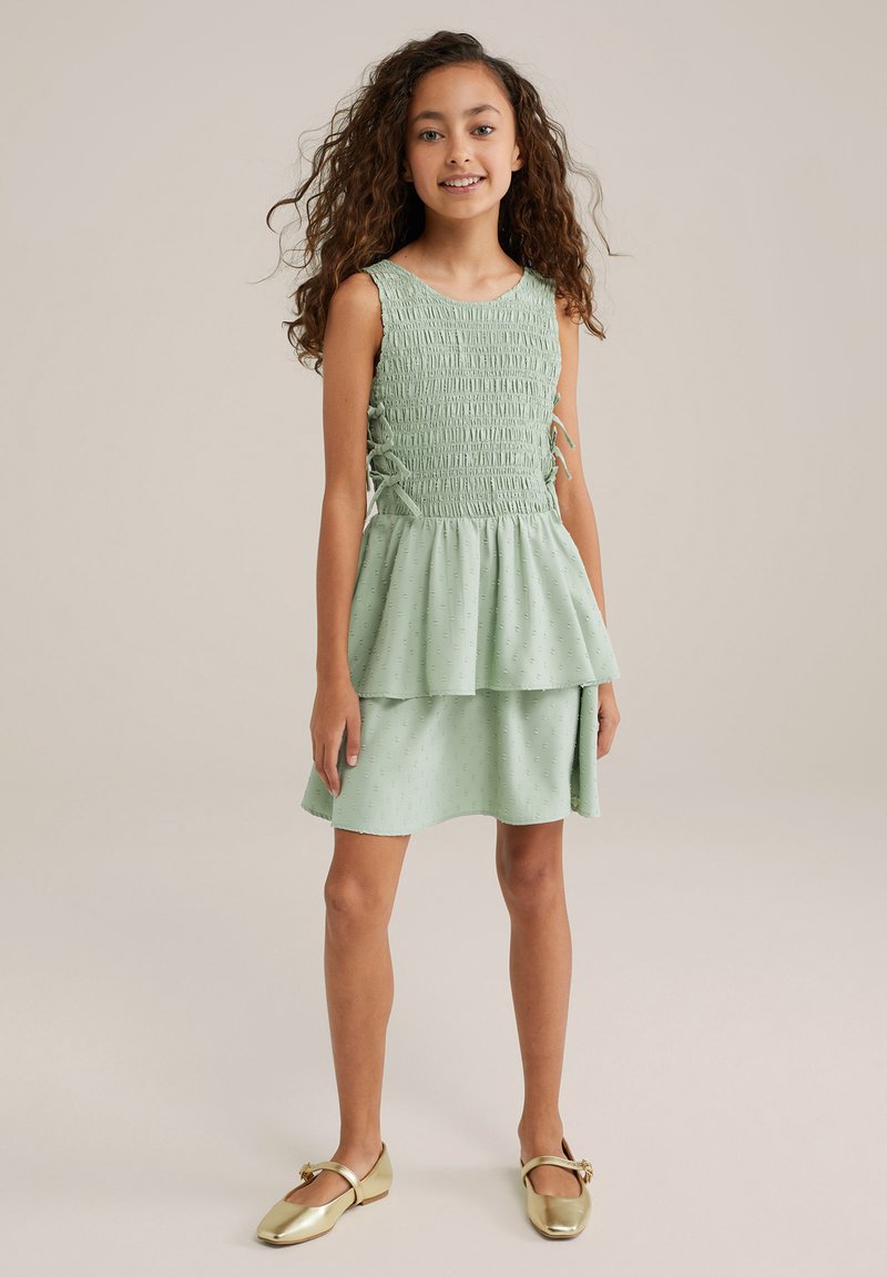 Mėtų žalios spalvos be rankovių suknelė su smock'd detalėmis, sluoksniuota sijonu ir tekstūruotu audiniu su subtiliais raštais. Dėvima su auksinėmis plokščiomis bateliais.