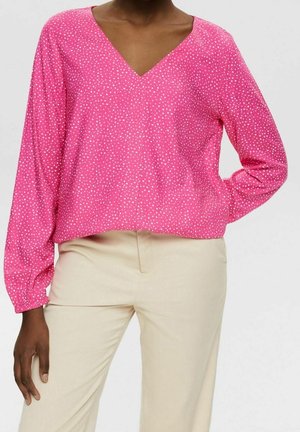 Personne portant un chemisier rose vif à manches longues avec col en V et des pois blancs, et un pantalon beige clair, la main reposant sur la hanche.