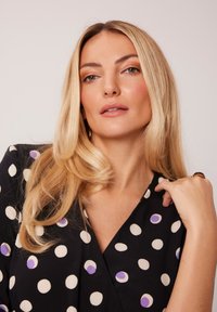 Czarna sukienka o kroju kopertowym z białymi i lawendowymi kropkami, z głębokim dekoltem i miękkiego materiału. Modelka prezentuje luźne, falowane blond włosy.