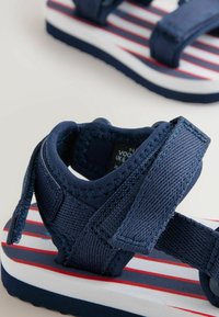 Sandale en tissu bleu marine avec des brides réglables à boucle auto-agrippante et une semelle blanche arborant des rayures horizontales rouges et bleues.