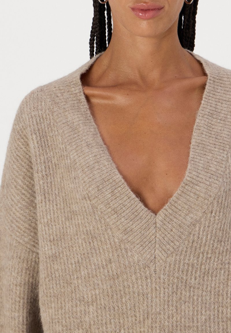 Beiger, gestrickter Pullover mit tiefem V-Ausschnitt und geripptem Muster. Der Stoff wirkt weich und gemütlich und weist dezente Farbvariationen auf.