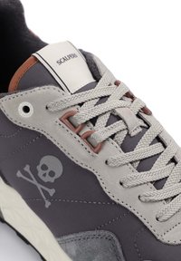 Adidași gri cu un superior din suede și material textil, având un logo cu schelet și oase încrucișate, șireturi gri și o guler și limbă cu umplutură.