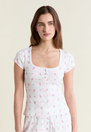 Młoda kobieta z długimi brązowymi włosami, ubrana w biały prążkowany, kwiatowy top z guzikami i dopasowane spodnie, na jednolitym tle.