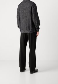 Veste noire structurée à fini texturé, portée sur un pantalon noir ample, avec un col et des poignets côtelés.