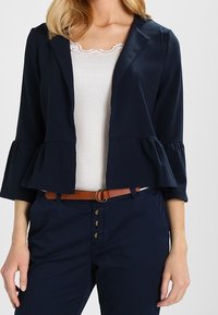 Mörkblå peplumblazer med trekvartslånga ärmar, texturerad tyg och öppen framtill. Bärs över en vit ribbad topp och mörkblå byxor, accessoriserad med ett brunt bälte.